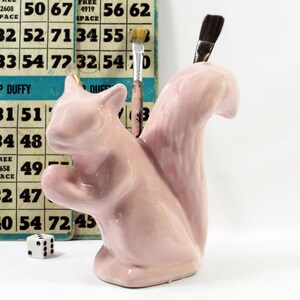 May include: A glossy light pink ceramic squirrel figurine designed as a desk organizer or pencil holder. The squirrel has a large, curved tail and a hollow opening at the top, currently holding two paintbrushes with wooden handles and dark bristles.