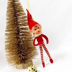 May include: A vintage Christmas elf ornament with a red suit, hat, and striped legs. The elf has a painted face and a white collar. A small gold-colored bottle brush tree and a die are in the background.