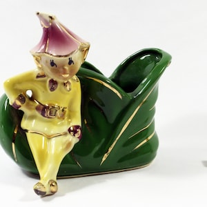 Puede incluir: Figura de cerámica vintage de un elfo apoyado en una jardinera verde en forma de hoja. El elfo viste un atuendo amarillo con detalles dorados y un sombrero rosa. Un dado con puntos negros está cerca.