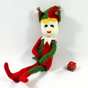 May include: A plush toy elf wearing a red and green jester hat with bells. The elf is sitting with its legs crossed and has a white face with black eyes and a red mouth. A red dice with gold dots is in the foreground.