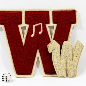 May include: A large, red felt letter "W" with a cream-colored border and a musical note. A smaller, cream-colored felt letter "W" is next to it. A white die with black dots is in the foreground. The items are likely for crafts or decoration.