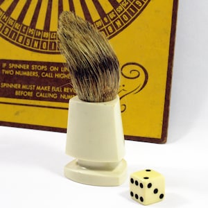 May include: A white ceramic shaving brush with a brown and white bristle brush head. A white dice with black dots is in the foreground.