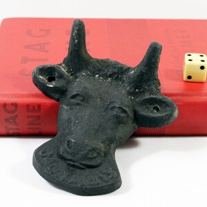 May include: A black cast iron cow head bottle opener. The opener is sitting on a red book with the word "STAG" visible on the cover.