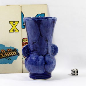 May include: A cobalt blue ceramic vase with two side handles. The vase has a bulbous base and a flared rim. The surface has a crackle glaze effect. A die and a vintage children's book are in the background.