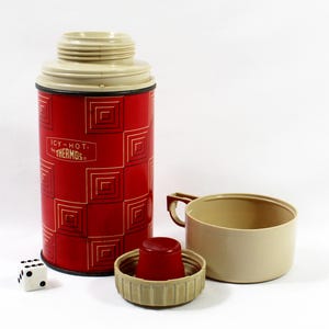 May include: A red and beige Thermos brand vacuum bottle with a geometric square pattern and the text Icy-Hot by Thermos. The set includes a beige screw-on cup with a handle, a beige lid with a red insert, and a small white six-sided die.