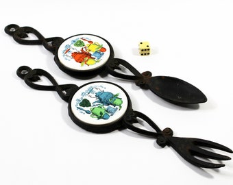 May include: A pair of black cast iron wall mounted spoon and fork holders with white ceramic inserts featuring colourful illustrations of kitchen utensils.