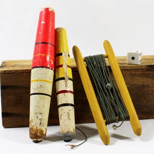 May include: Two cylindrical wooden fishing floats with chipped red, yellow, and white paint, alongside a wooden handline spool wrapped in dark green fishing line and a single white six-sided die with black pips, all arranged in a row.