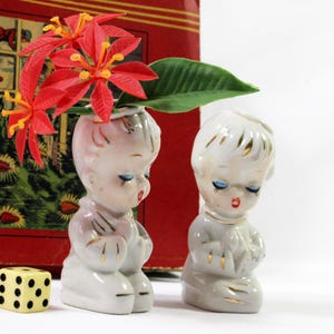 May include: Two ceramic figurines of children in prayer, one holding red flowers. The figurines are white with gold accents, closed eyes, and red lips. A yellow and black die is in the foreground. A red box with a vintage design is in the background.