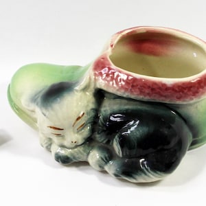 May include: A ceramic planter in the shape of a sleeping cat. The cat is white with black markings and closed eyes. The planter has a green base and a red-rimmed opening. A white die with black dots is next to the planter.
