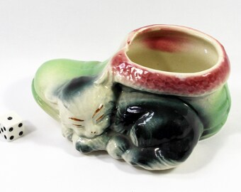 May include: A ceramic planter in the shape of a sleeping cat. The cat is white with black markings and closed eyes. The planter has a green base and a red-rimmed opening. A white die with black dots is next to the planter.