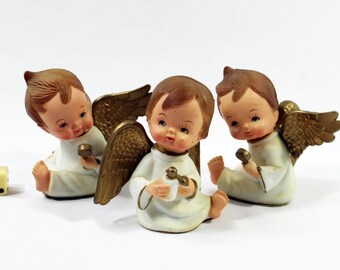 May include: Three small, white angel figurines with gold wings. Each angel is holding a small, gold musical instrument. The angels are sitting on a white surface.