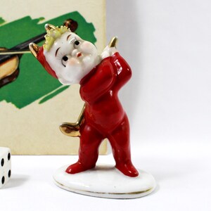 May include: A small, red ceramic figurine of a devil-like character with a white face and gold accents, holding a golf club. The figurine is on a white base with a gold rim. A die and a vintage card with a violin illustration are in the background.