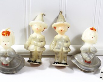 May include: Four vintage pilgrim-themed candles, two are boys wearing hats and two are girls with bonnets. The candles are white with gray and red accents.