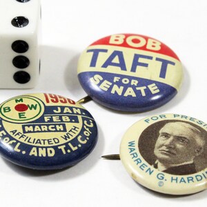 May include: Three vintage political campaign buttons. The top button is blue, white, and red with the text "Bob Taft for Senate". The bottom left button is blue and white with the text "A.F.OfL. and T.L.C.O. Affiliated with 1950 Jan. Feb. March". The bottom right button is white with the text "For President Warren G. Harding".
