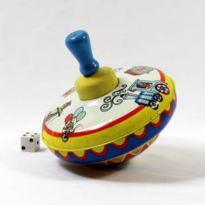 May include: A vintage metal spinning top toy featuring a blue handle and a yellow base. The body is decorated with colorful illustrations of a train, a figure with balloons, and geometric patterns. A small white six-sided die sits nearby.