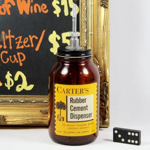 May include: A vintage brown glass jar labeled "CARTER'S Rubber Cement Dispenser" with a metal dispenser top. A black domino is on the right. The background includes a chalkboard with handwritten text and a gold-framed mirror.