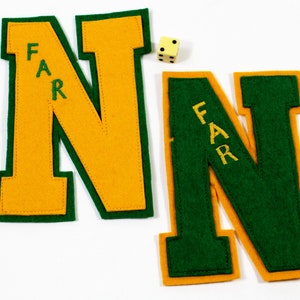 May include: Two felt letter patches in the shape of the letter N. The letter on the left is yellow with a green border. The letter on the right is green with a yellow border. Both letters have the word "FAR" embroidered on them in yellow thread.