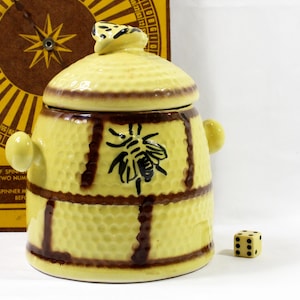 May include: A yellow ceramic jar with a lid, featuring a bee design and brown accents. The jar has a textured surface and small handles. A die with black dots sits nearby. A vintage game board is in the background.