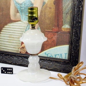 May include: A vintage white glass table lamp with a brass-colored socket and a decorative base. The lamp is next to a framed vintage print with a black frame. A domino is on the surface.