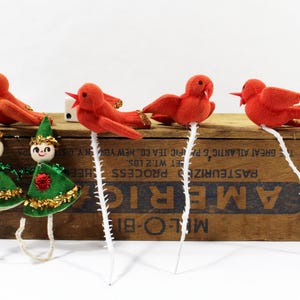 Puede incluir: Una colección de adornos navideños vintage. La imagen presenta cuatro pájaros rojos de fieltro sobre tallos blancos y dos figuras de duendes con trajes de terciopelo verde y adornos dorados. Los adornos están colocados sobre una caja de madera.