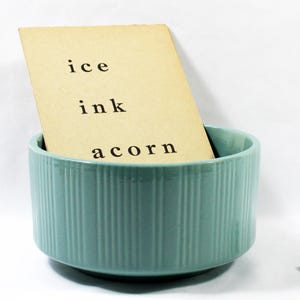 May include: A light teal ceramic bowl with vertical ridges holds a tan card with the words "ice," "ink," and "acorn" printed on it. A white die with black dots sits to the right. The items are on a white background.