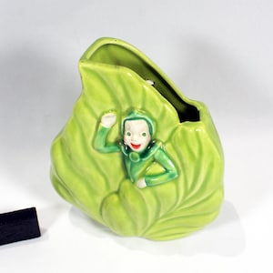 May include: A light green ceramic vase shaped like a large leaf with a small figure emerging from the center. The figure has a green hood, a pale face, and a red mouth. A small black rectangular object is positioned to the left of the vase.