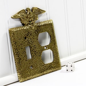 May include: A brass electrical outlet cover with a textured surface and an eagle emblem. The cover has openings for a light switch and two electrical outlets. A white die with black dots is next to the cover.
