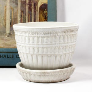 May include: Off-white ceramic planter with a matching saucer. The planter has a textured, embossed design with vertical and horizontal patterns. The saucer is round and has a similar embossed pattern. The planter is suitable for indoor or outdoor use.
