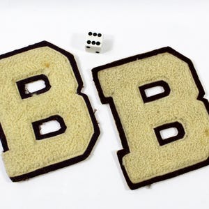 May include: Two vintage beige felt letter "B" patches with dark brown borders. A white die with black dots is positioned above the patches. The patches have a textured, fuzzy appearance.