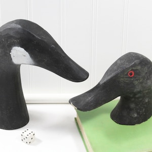May include: Two carved wooden goose heads, one black with a white stripe and the other black with red eyes. The larger goose head is on a white surface and the smaller goose head is on a green book.