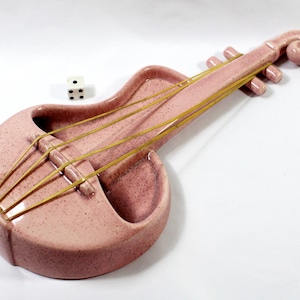 May include: A pink ceramic violin-shaped ashtray with yellow strings and a curled scroll. A white die with black dots sits nearby. The ashtray has a speckled finish and a unique, artistic design.