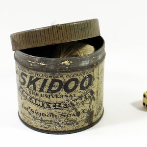 May include: A vintage tin can with a yellow label that reads "Skidoo The Universal Health Cleanser The Skidoo Soap Co.  Columbu Ohio". The lid is open and a ball of string is inside the can. A white dice with black dots is on a white surface.