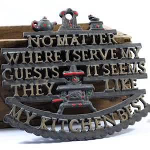 May include: A dark gray cast iron trivet featuring raised lettering that reads, "NO MATTER WHERE I SERVE MY GUESTS IT SEEMS LIKE MY KITCHEN BEST." The design includes small, colorful painted accents on a teapot, a stove, and a coffee grinder motif.