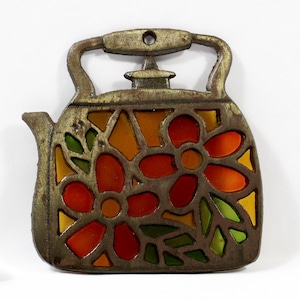 May include: A decorative metal teapot-shaped trivet with a stained glass design. The trivet features a floral pattern in shades of red, orange, yellow, and green. The handle and body have a distressed, antique finish.
