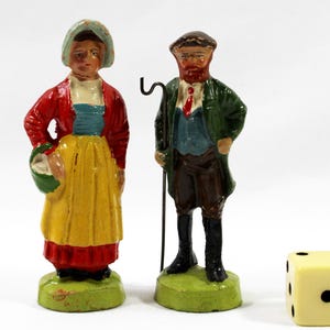 May include: A pair of vintage painted figurines depicting a woman in a red jacket and yellow skirt, and a man with a beard in a green coat. A yellow die with black dots is also present.