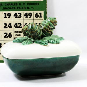May include: A ceramic trinket box with a white lid and a green base. The lid is adorned with green pinecone-shaped decorations. The box is next to a bingo card and a die.