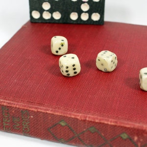 May include: Four white dice with black dots on a red book cover. The dice are arranged in a line, with the first die showing a four, the second die showing a five, the third die showing a six, and the fourth die showing a three.