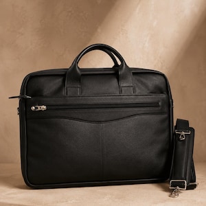 May include: A black leather briefcase featuring two top handles, a horizontal zippered front pocket with a silver-toned metal pull, and a detachable shoulder strap with silver-toned metal clips attached to the side of the rectangular bag.