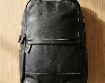 May include: A black leather backpack featuring a rounded top, a front zippered pocket with a leather pull tab, and side pockets. The bag is accented with visible white contrast stitching along the seams and includes a top handle for carrying.
