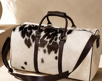 May include: A duffel bag featuring a white and dark brown cowhide pattern with dark brown leather handles, trim, and an adjustable shoulder strap. The bag has a top zipper closure and metal hardware, presenting a cylindrical shape for travel.