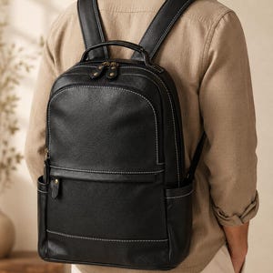 May include: A black backpack with a textured finish and white contrast stitching. It features a main zippered compartment, a smaller front zippered pocket with a brass-toned snap button detail, two side pockets, and adjustable shoulder straps.