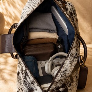 May include: A duffel bag with a black and white cowhide pattern and dark brown leather handles. The interior is lined with dark blue fabric and contains folded clothing, a brown leather pouch, a bottle, headphones, and a magazine with the text CEREAL.