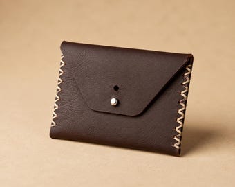 May include: A dark brown leather card holder with a triangular flap closure. The edges are secured with visible light-colored cross-stitching. A single silver-toned metal stud fastener is centered on the front flap to keep the wallet closed.