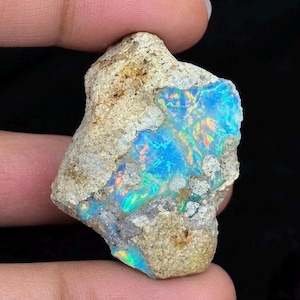 May include: A raw, unpolished opal specimen featuring a rough, beige, and sandy-textured rock matrix embedded with vibrant, iridescent patches of bright blue, green, and orange flashes that shimmer across the irregular surface of the stone.