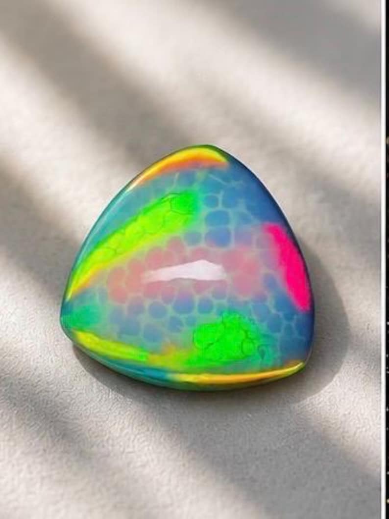 May include: A triangular cabochon gemstone featuring a translucent blue base with a honeycomb-like pattern. The surface displays vibrant, iridescent flashes of neon green, bright pink, and yellow, creating a multi-colored play of light.