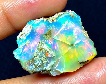 May include: A raw, unpolished opal gemstone displaying vibrant flashes of neon blue, green, and yellow light. The stone features a translucent, crystalline structure with irregular, jagged edges and patches of rough, earthy, light-colored matrix.