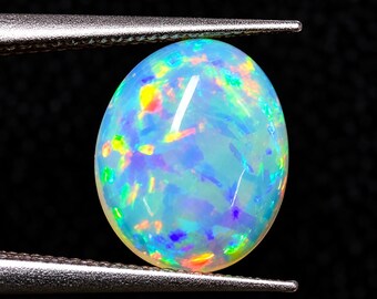 May include: An oval-shaped cabochon opal gemstone featuring a translucent light blue base with vibrant flashes of yellow, orange, green, and purple play-of-color. The gemstone is held securely between the metal tips of a pair of tweezers.