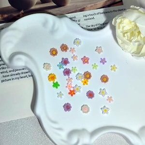 以下が含まれることがあります： ピンク、黄色、青、紫、オレンジ、白の色合いを含む、さまざまな形とサイズの小さな多色の樹脂製フラワーカボションのコレクション。大きな白い花が添えられた、縁が波打つ白いトレイの上に散らばっています。