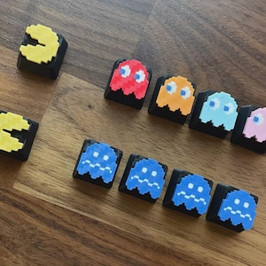 May include: A collection of eleven black mechanical keyboard keycaps featuring pixelated designs. Two yellow Pac-Man characters, one red, one orange, one light blue, one pink, and five dark blue ghost characters are attached to the keycaps.