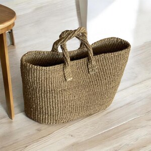 May include: A tan woven basket bag with a structured oval base and two braided handles. The bag features a dense, textured weave pattern throughout its body, creating a sturdy and open-top design for carrying items. The natural fiber material has a light brown color.